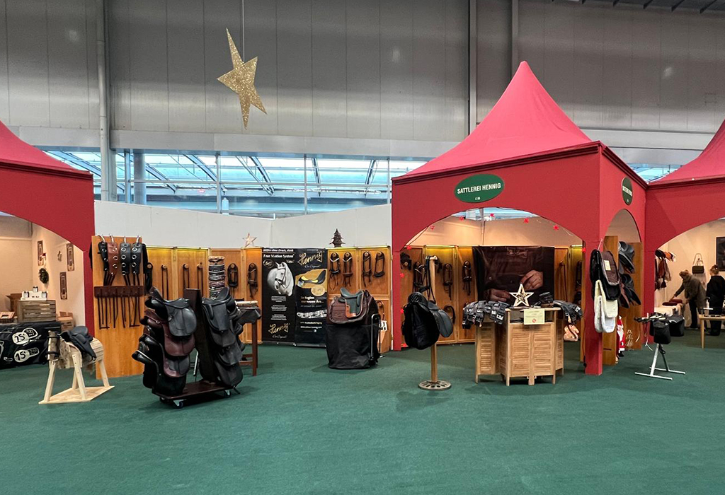 Auf dem Bild sieht man den großen Messestand der Sattlerei Hennig. Man sieht eine Auswahl an Mustersätteln, Zaumzeugen, auch ein Holzpferd mit einem Kindersattel ist im Vordergrund zu sehen. Die Messe ist schön arrangiert, mit roten Bogenpavillons, grünen Teppichen und einem Weihnachtsstern.